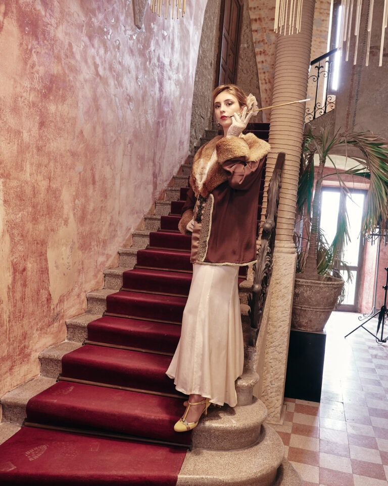 Woman in a vintage fur coat and cream skirt poses on a red-carpet staircase in a grand, textured interior.