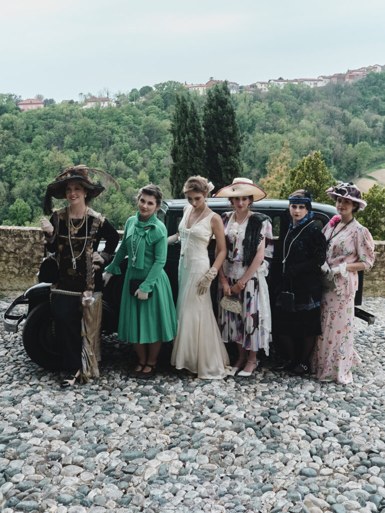 Six women in vintage dresses and hats pose beside a dark car on a cobblestone path with green hills in the background.